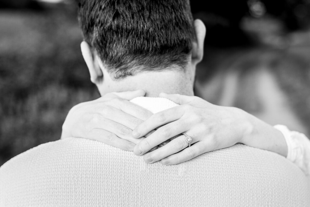 A black-and-white photo showing the back of a man's head and shoulders as a woman’s hands, with an engagement ring on her finger, rest gently on his neck.