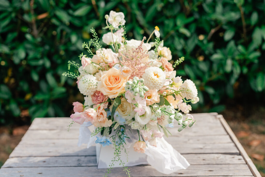 A pastel-colored floral arrangement with roses, peonies, and greenery sits on a rustic wooden table outdoors, with leafy green bushes in the background.