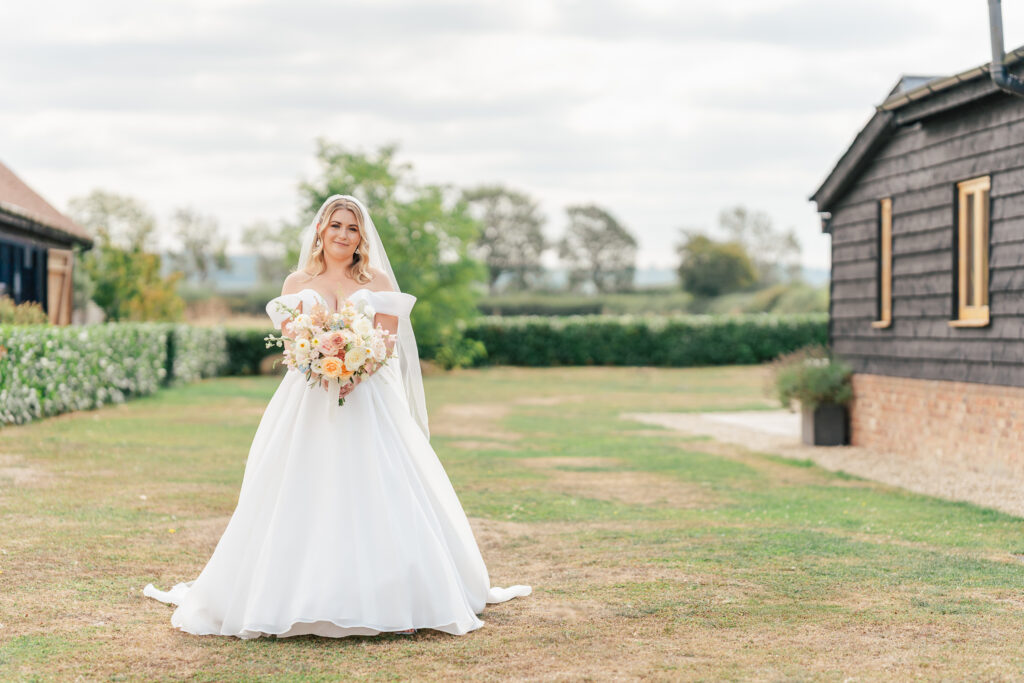 A bride in a white wedding dress holds a bouquet of flowers outdoors on a grassy lawn. She stands between rustic buildings with trees and a cloudy sky in the background.