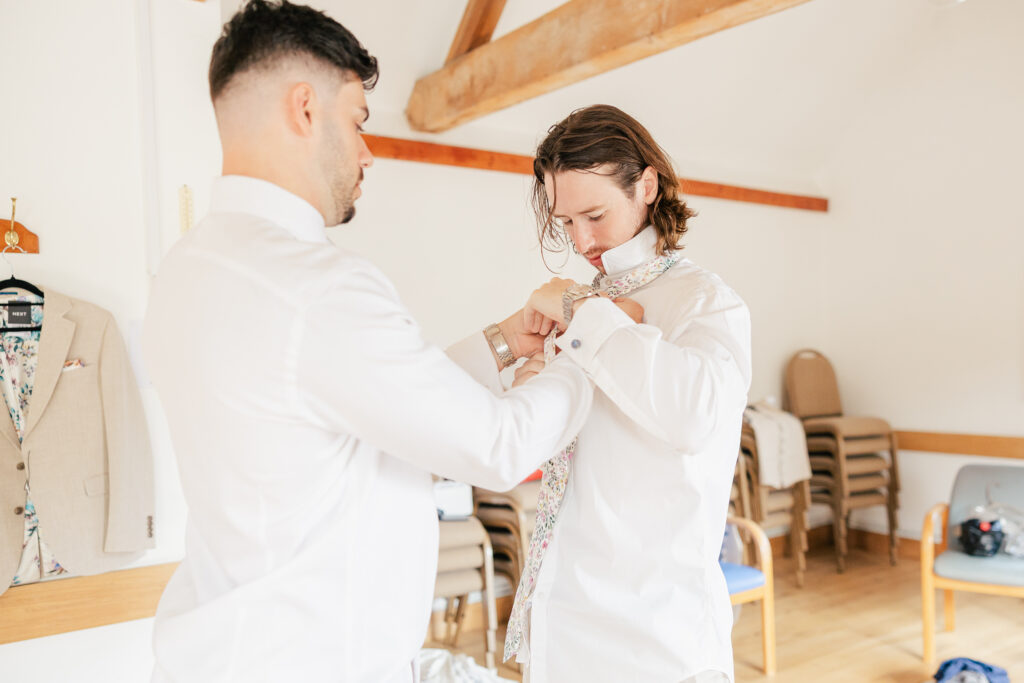 Two men in white shirts stand in a bright room; one helps the other adjust his floral tie. Stacked chairs and a beige suit jacket are visible in the background. The scene suggests preparation for a formal event.