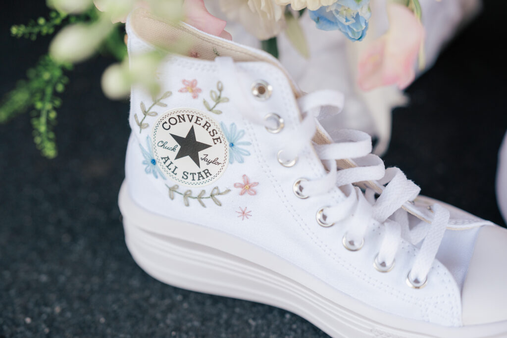 A close-up of a white Converse All Star high-top sneaker with pastel floral embroidery around the logo, worn by someone on a dark textured surface. Some blurred white and green flowers are visible in the background.