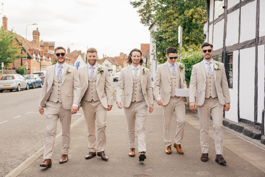 Five men in matching light beige suits with floral ties walk confidently down a sidewalk in a town, smiling. They appear to be part of a wedding party, likely groomsmen, on a sunny day.