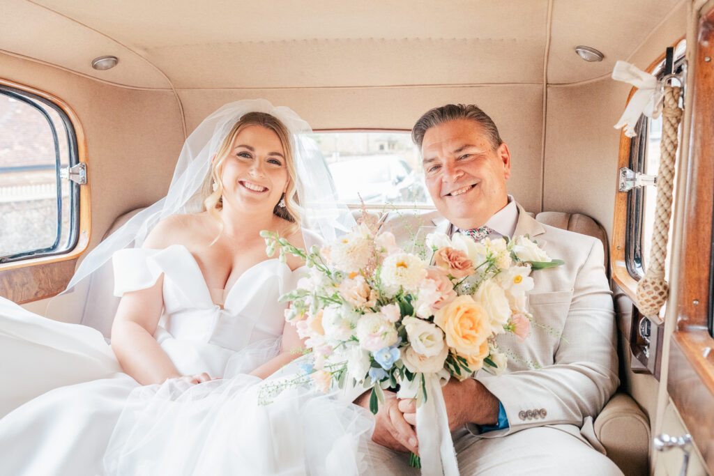 A smiling bride in a white gown and veil sits in the back of a vintage car next to a man in a light suit, both holding a large bouquet of pastel flowers.