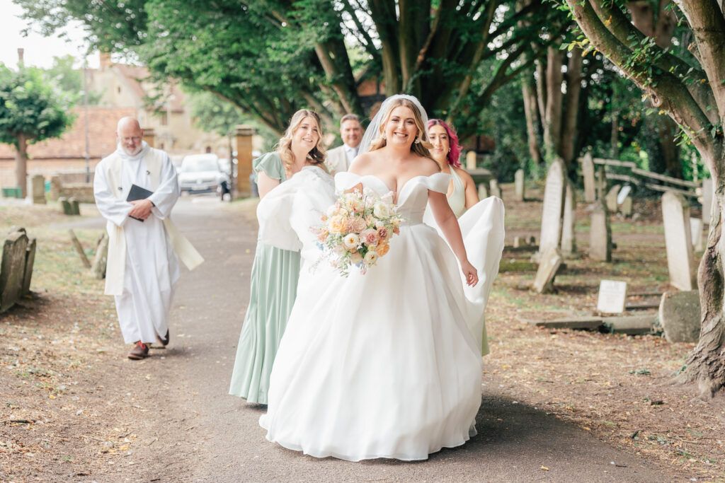 A smiling bride in a white gown holds a bouquet while walking outside on a tree-lined path, followed by bridesmaids and a clergyman. The group appears joyful, surrounded by greenery and gravestones.