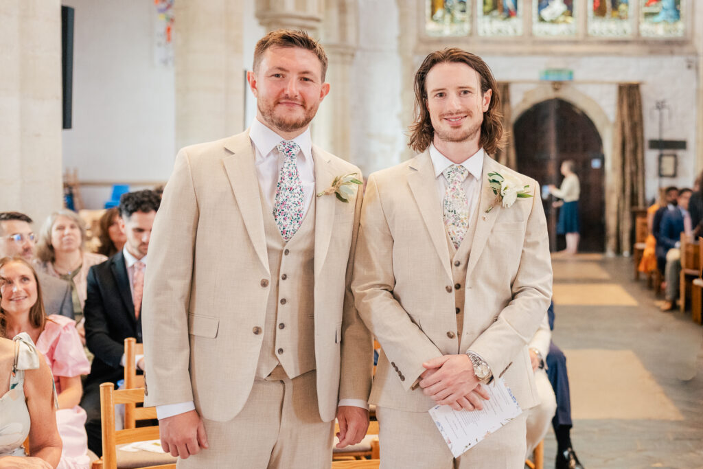 Two men in light beige suits with floral ties stand side by side in a church, smiling at the camera. People are seated in the background, and stained glass windows are visible behind them.