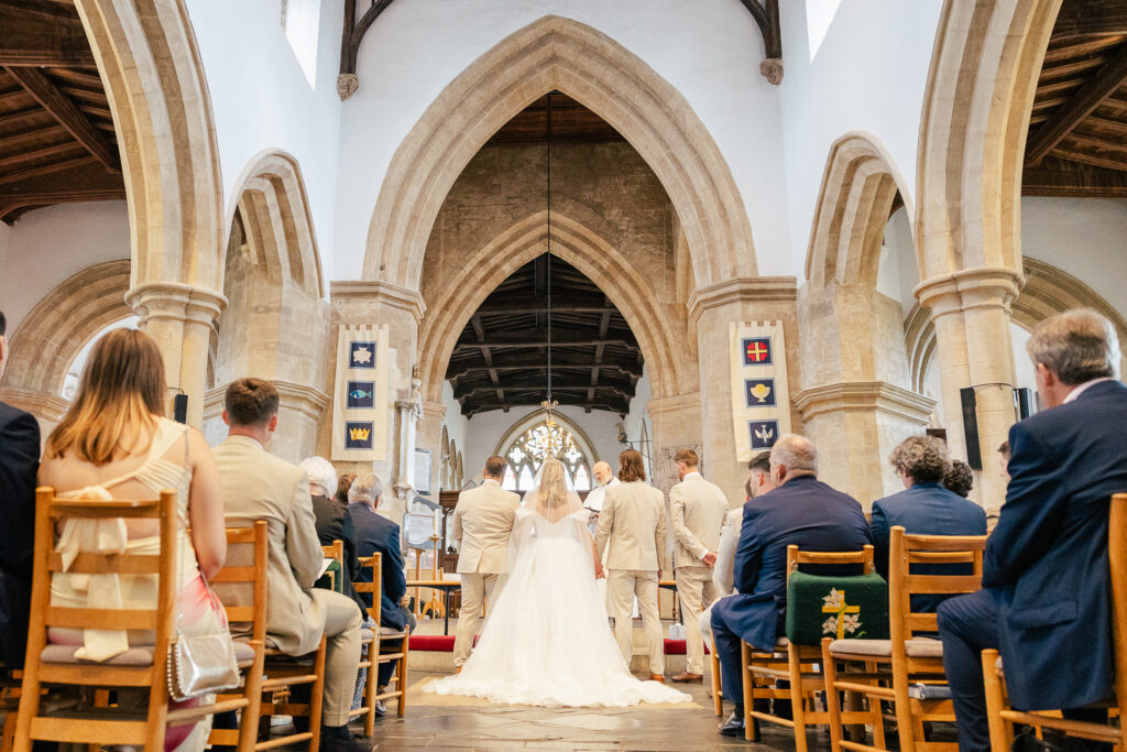 A bride and groom stand at the altar in a grand church, surrounded by their bridal party and seated guests. The church features high arches, wooden ceilings, and stained glass windows, creating a bright, solemn atmosphere.