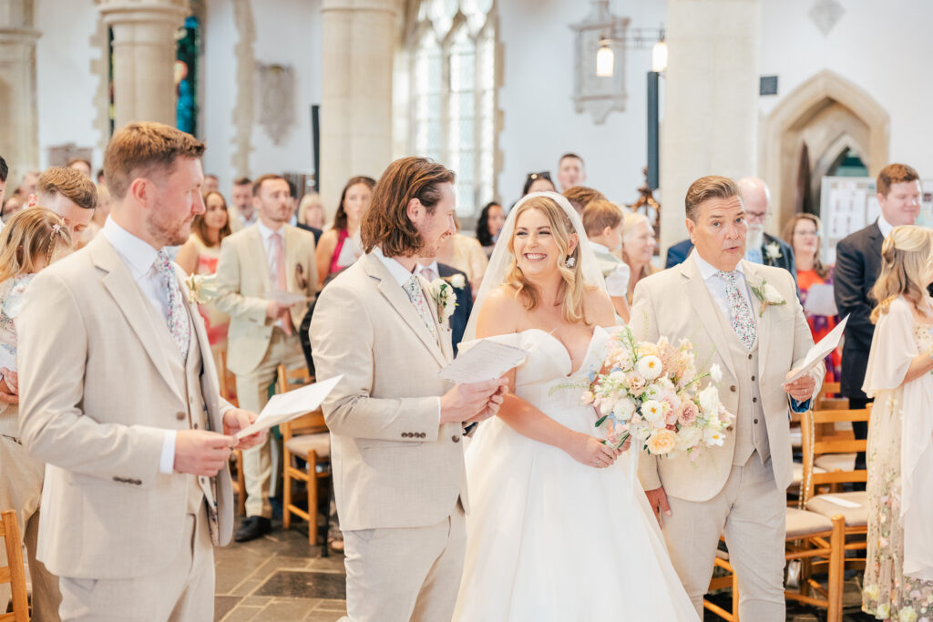 A bride in a white dress smiles while standing between two men in light-colored suits, holding a bouquet of flowers inside a bright, decorated church filled with well-dressed guests.