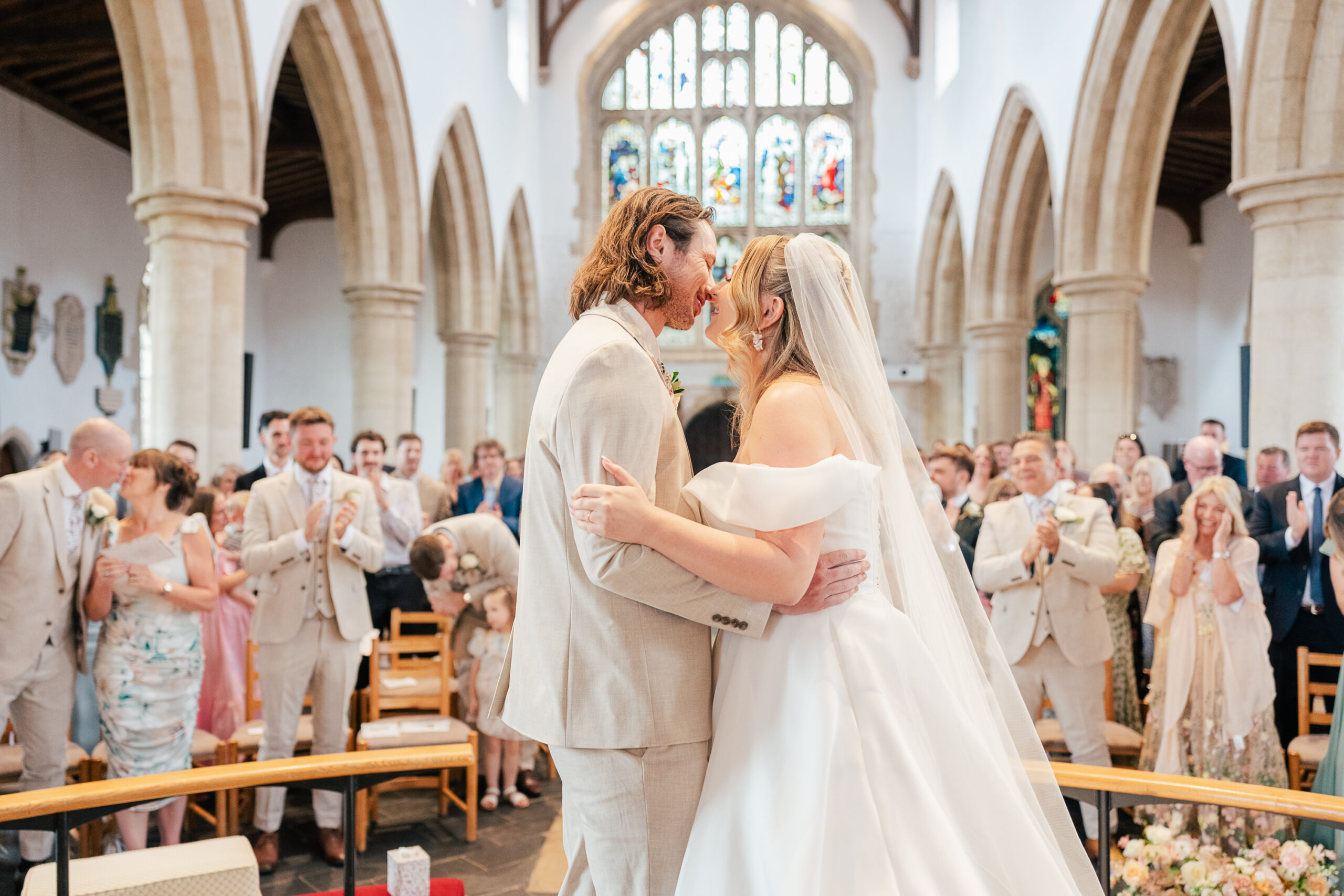 A bride and groom share a kiss at the altar in a church filled with cheering guests, bathed in natural light from grand arched windows. The bride wears a white dress and veil; the groom is in a light suit.