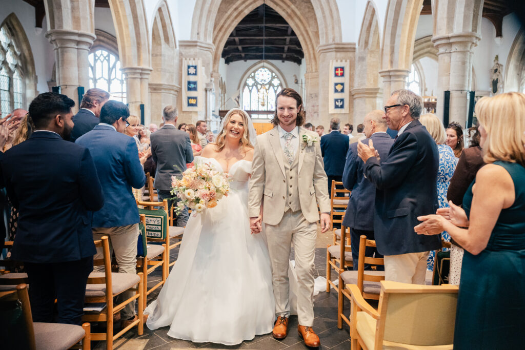 A bride in a white dress and a groom in a beige suit walk down the aisle, smiling and holding hands, as guests in formal attire stand and applaud inside a bright church.