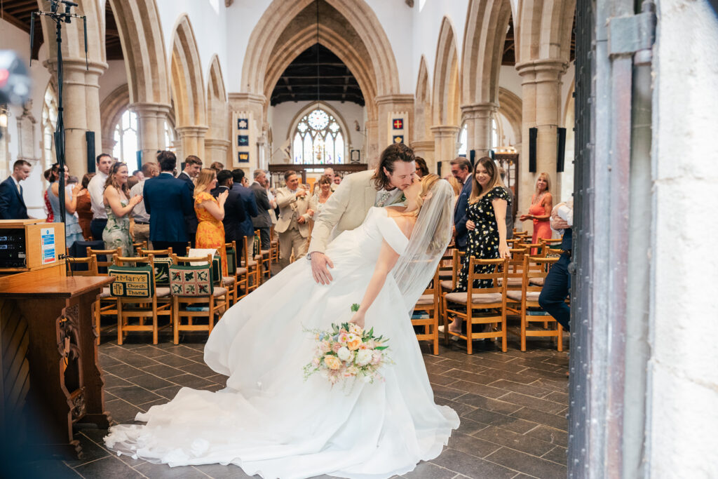 A bride and groom share a joyful dip and kiss in the aisle of a church, surrounded by smiling guests. The bride holds a bouquet of flowers, and sunlight streams through the arched windows behind them.