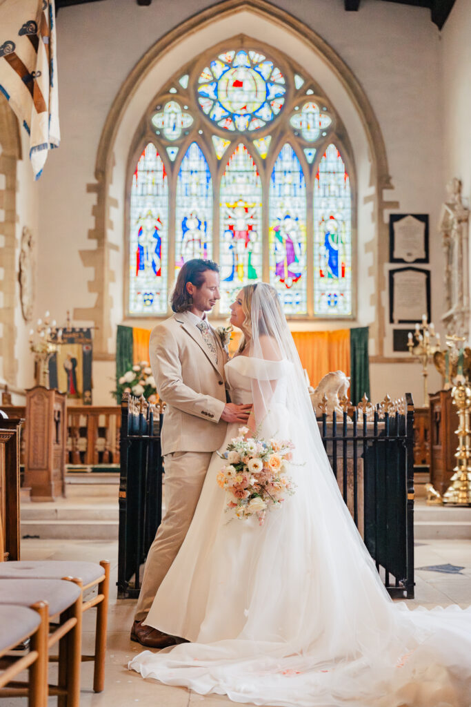 A bride and groom stand close together in a church, gazing into each other’s eyes. The bride holds a bouquet and wears a long white dress with a veil; the groom wears a beige suit. Stained glass windows shine behind them.