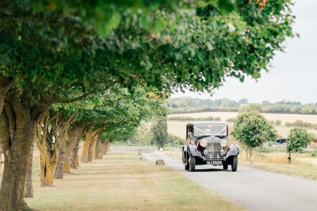 A vintage car drives down a narrow rural road lined with green trees, with open fields and rolling hills in the background under a partly cloudy sky.