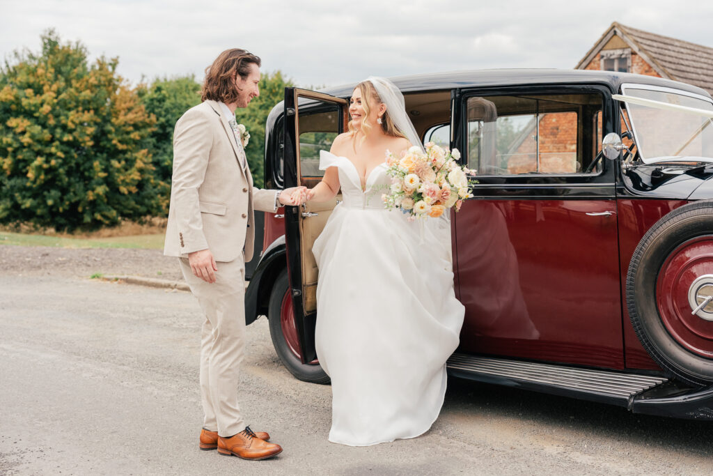 A bride in a white gown with a bouquet steps out of a vintage maroon car, smiling at a groom in a beige suit who is holding her hand. They are outdoors near greenery and a rustic building.