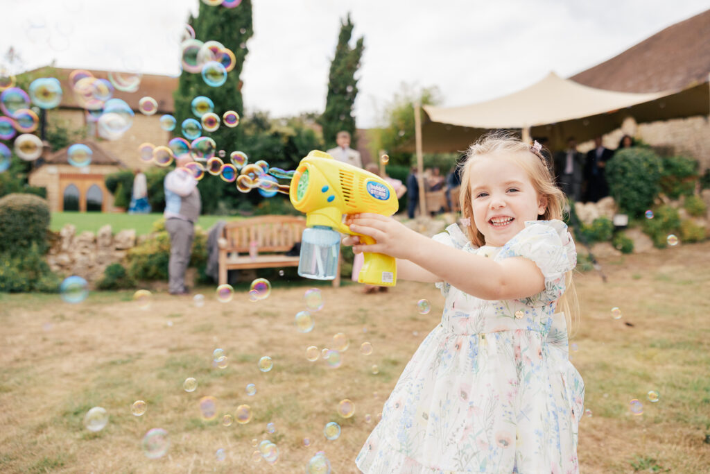 A young girl in a floral dress smiles while playing with a yellow bubble gun outdoors. Soap bubbles float around her, and several people are in the background near a tent and greenery.