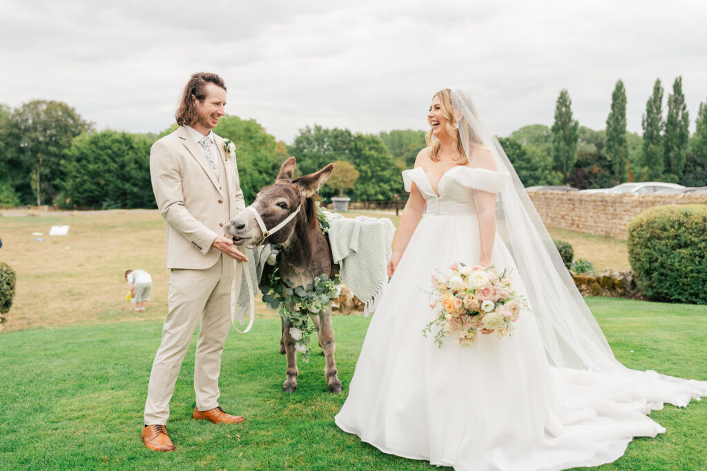 A bride in a white gown and a groom in a beige suit stand on grass, happily posing with a donkey adorned with greenery and a blanket, with a bouquet in the bride's hand and trees in the background.
