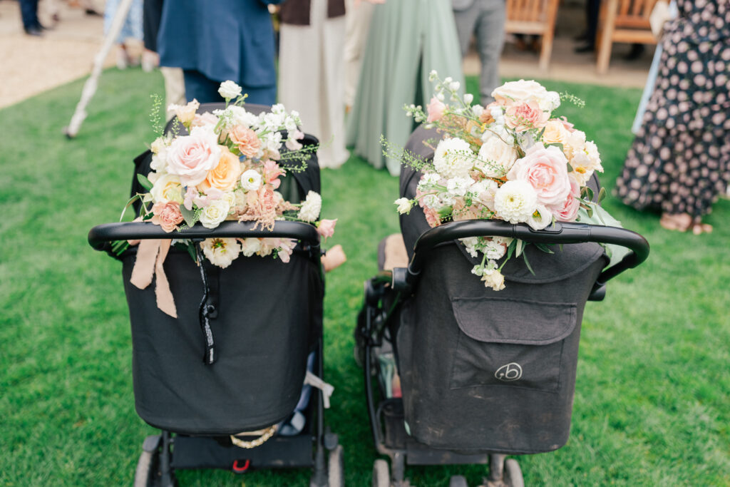 Two black strollers decorated with pastel floral bouquets sit side by side on green grass at an outdoor event with people standing in the background.