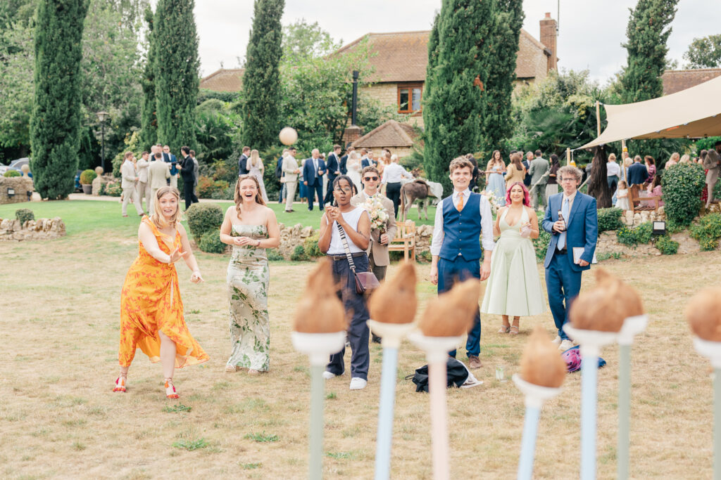 A group of well-dressed people outdoors play a lawn game, aiming to toss balls at targets on sticks. The scene is festive, with greenery, a tent, and a rustic house in the background.