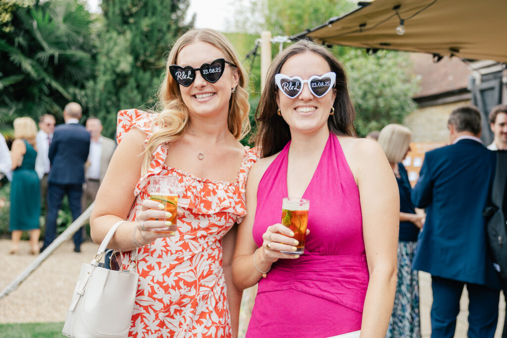 Two women at an outdoor event smile at the camera, holding drinks. One wears a pink floral dress, the other a magenta halter dress. Both wear heart-shaped sunglasses that say "Rob & Rose." People and greenery are in the background.