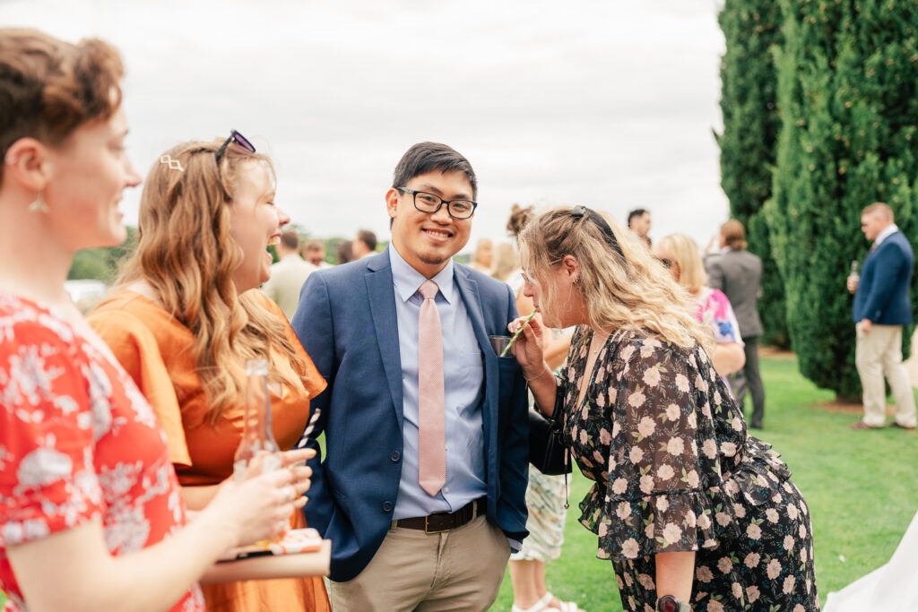 Four people stand together outdoors, dressed in semi-formal attire, smiling and laughing. One woman in a floral dress drinks from a glass, while others hold drinks, with trees and more people in the background.