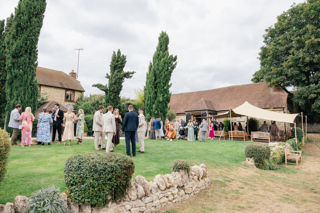A group of people dressed in formal attire gather and mingle on a lawn outside rustic buildings and a marquee tent, surrounded by trees and stone walls on a cloudy day.