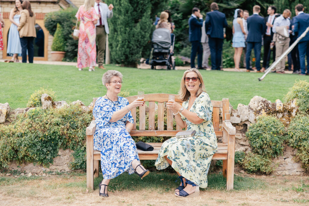 Two women in floral dresses sit on a wooden bench outdoors, smiling and raising glasses of champagne. A group of people socializes in the background on a grassy area. It appears to be a celebratory event or gathering.