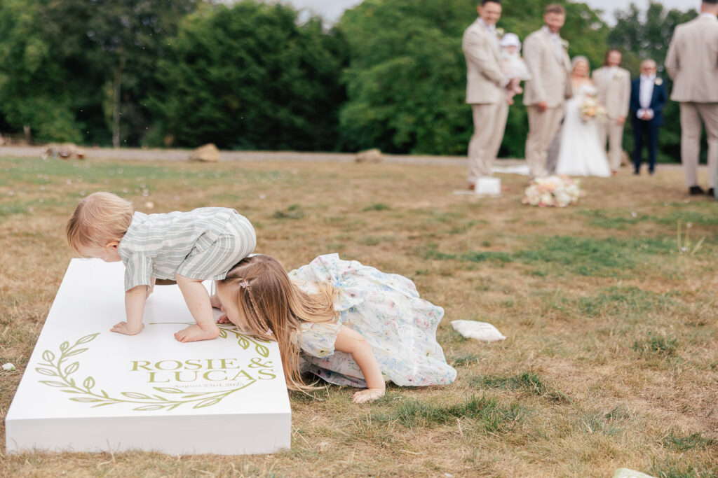 Two young children play on a white cornhole board labeled "Rosie & Lucas," while wedding guests in pale suits and a bride stand blurred in the background on a grassy lawn.