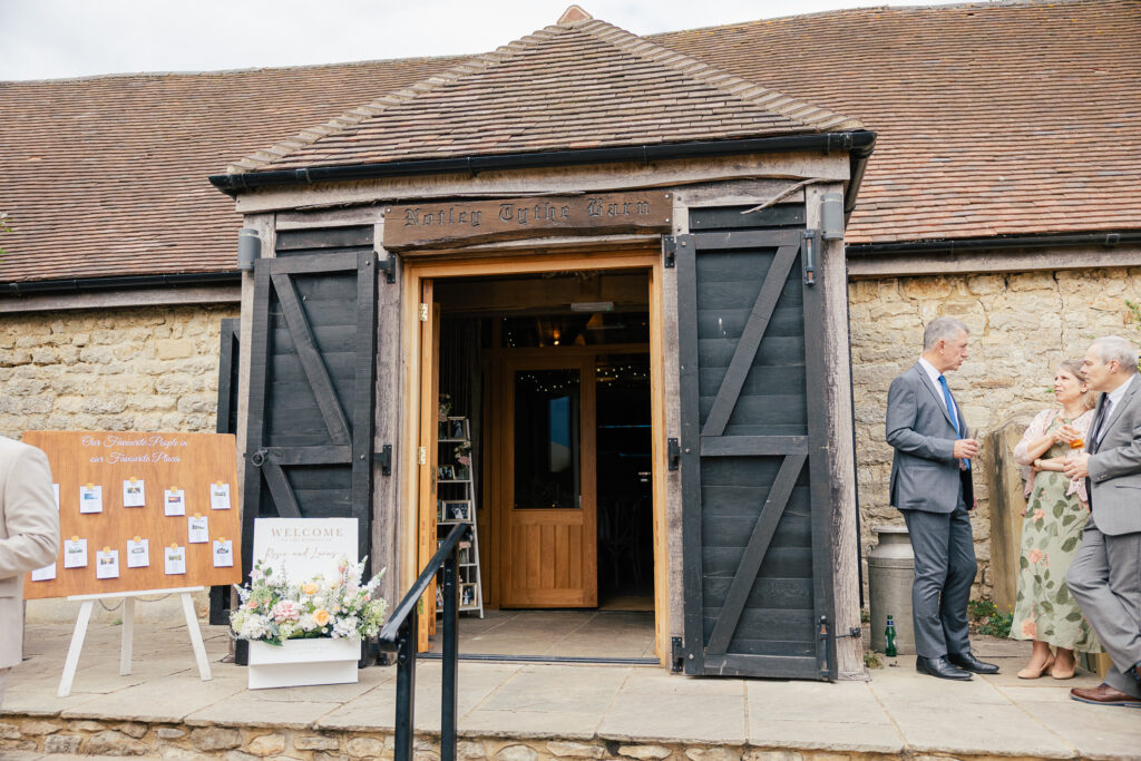 A rustic barn entrance with open wooden doors, a welcome sign with flowers, and a seating chart displayed on a board. Three well-dressed people are talking near the entrance on a stone patio.