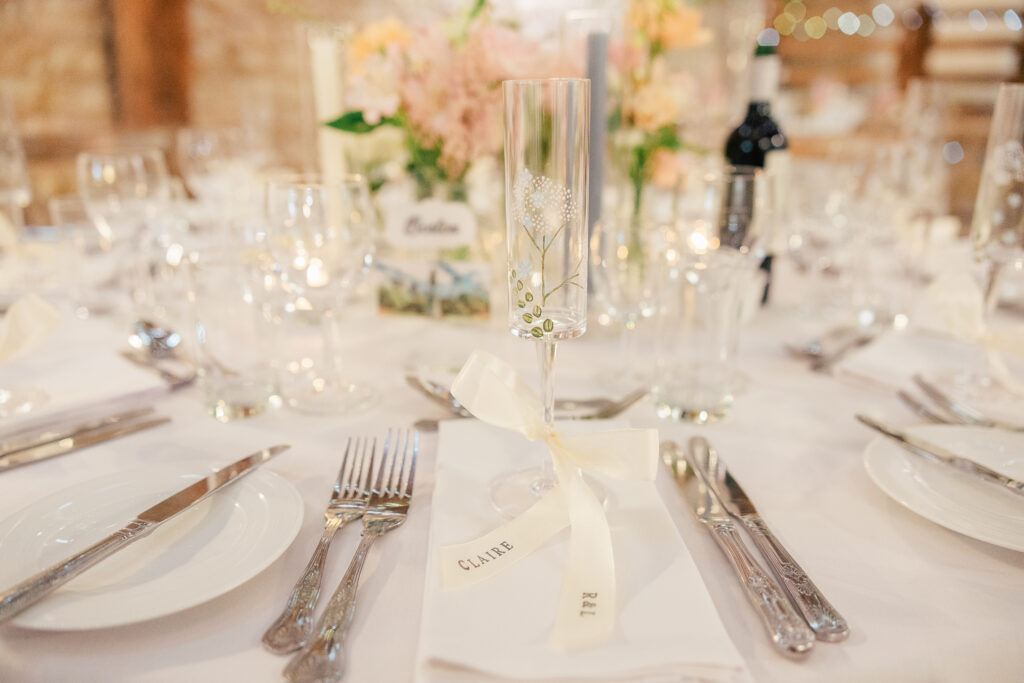 A close-up of an elegant wedding table setting with a personalized champagne flute, white napkin, and a ribbon. The table is decorated with flowers, wine, candles, and neatly arranged cutlery and glassware.