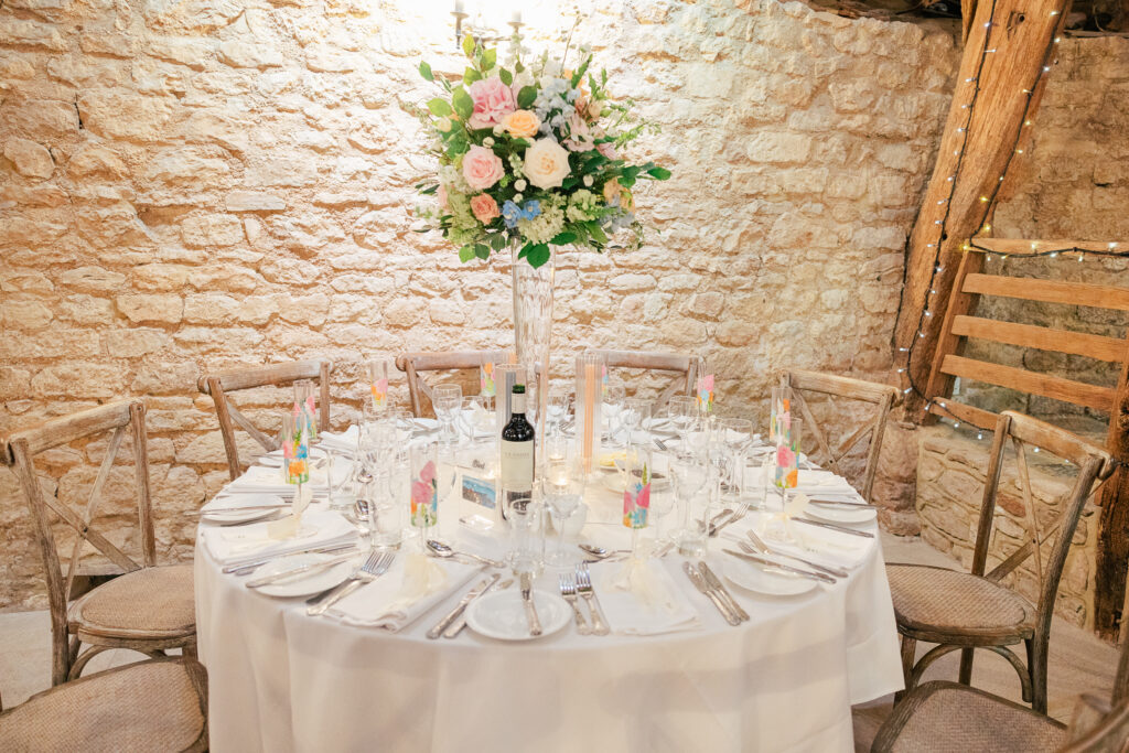 A round table set for a formal event with a white tablecloth, glasses, cutlery, and napkins. A tall floral centerpiece with pink and white flowers stands in the middle. Rustic wooden chairs and a stone wall are in the background.