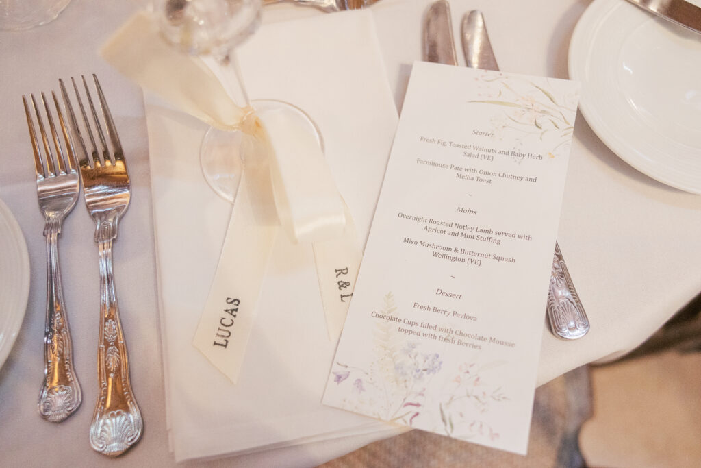 A formal place setting with silver cutlery, a white napkin tied with a ribbon labeled "LUCAS," and a printed menu listing starter, main, and dessert options on a white tablecloth.