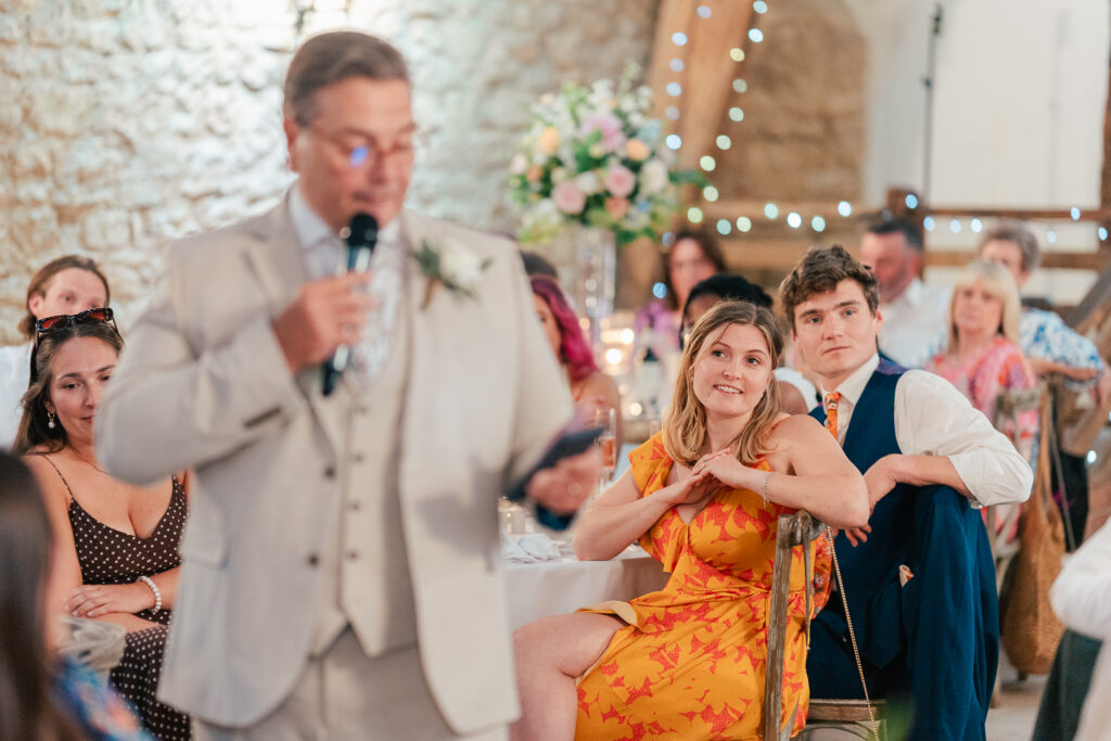 A man in a light suit speaks into a microphone at a formal event, while guests seated at decorated tables listen attentively. A woman in a yellow dress and a man in a dark vest are in focus, watching the speaker.