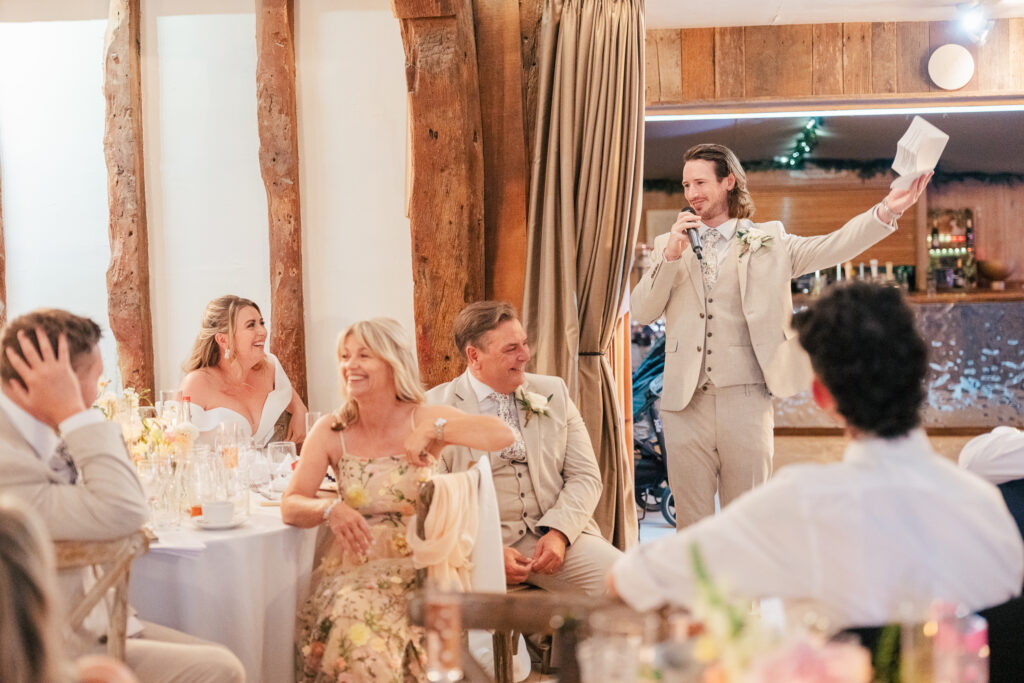 A man in a light suit gives a speech with a microphone at a wedding reception. Guests, including a smiling bride, groom, and others at their table, listen and laugh in a warmly lit rustic venue.