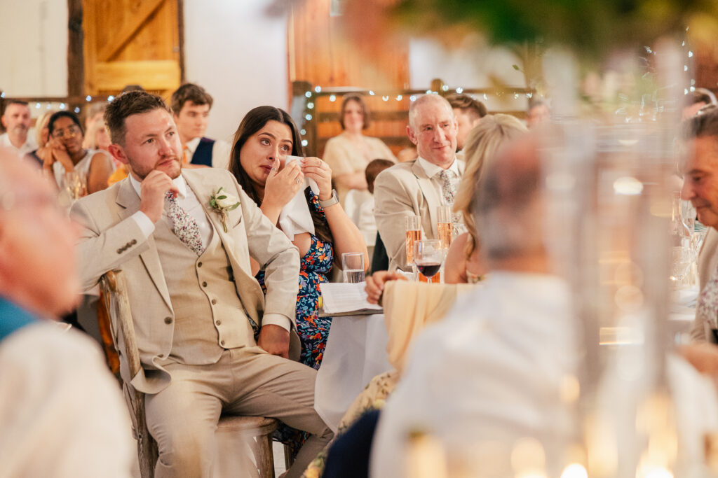 A man in a beige suit and a woman in a floral dress sit among wedding guests, both wiping away tears, surrounded by people attentively watching an unseen event, in a warmly lit, decorated venue.