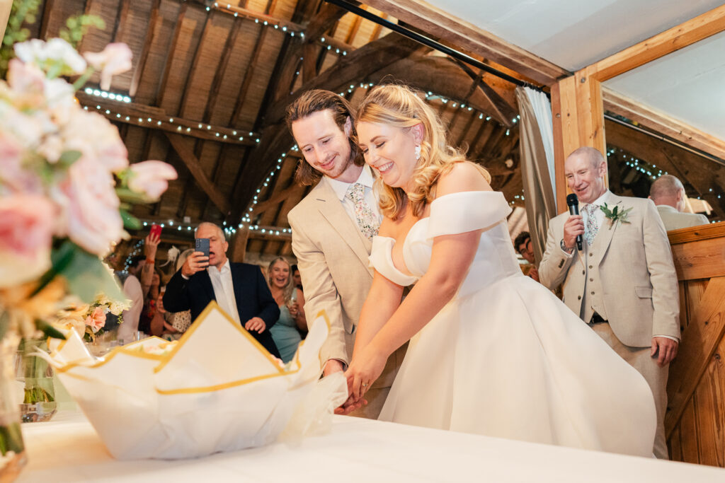 A bride and groom, both smiling, cut their wedding cake together in a warmly lit rustic venue. Guests watch and take photos in the background, and a man with a microphone stands nearby. Flowers are in the foreground.
