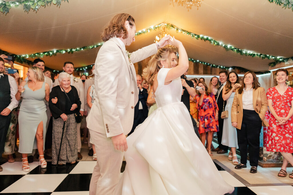 A bride and groom dance joyfully on a black-and-white checkered floor, surrounded by smiling guests. The bride twirls in her white dress under a tent decorated with greenery and string lights.