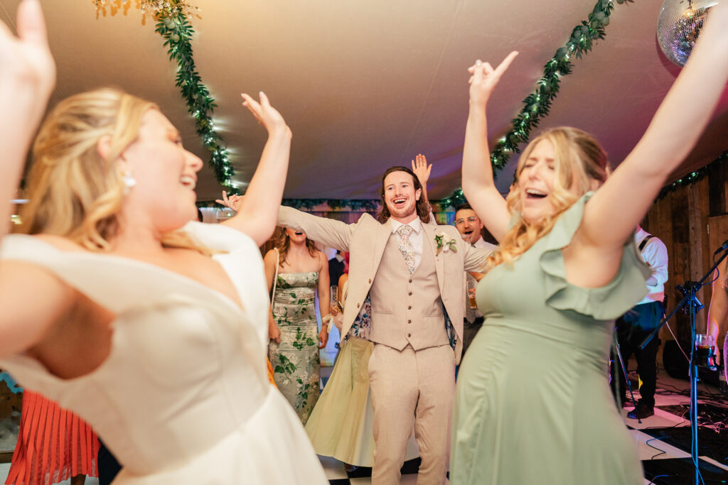 A group of people dressed in formal attire joyfully dance and raise their arms at a lively indoor celebration decorated with greenery and a disco ball.