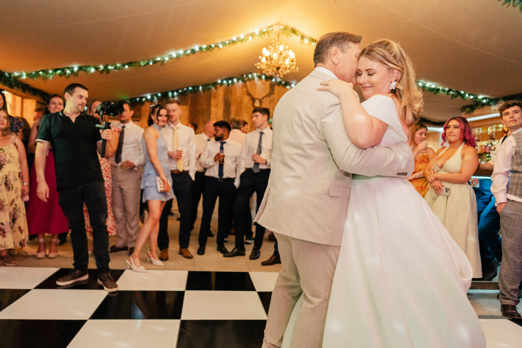 A bride in a white dress and a groom in a light suit share a dance on a black-and-white checkered floor, surrounded by smiling guests dressed in formal attire under a warmly lit, decorated tent.