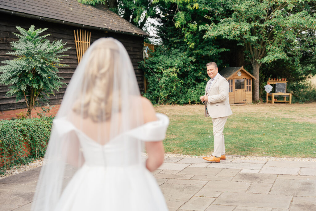 A bride in a white dress and veil walks toward a smiling groom in a light suit outside, with greenery, a wooden shed, and a small playhouse in the background.