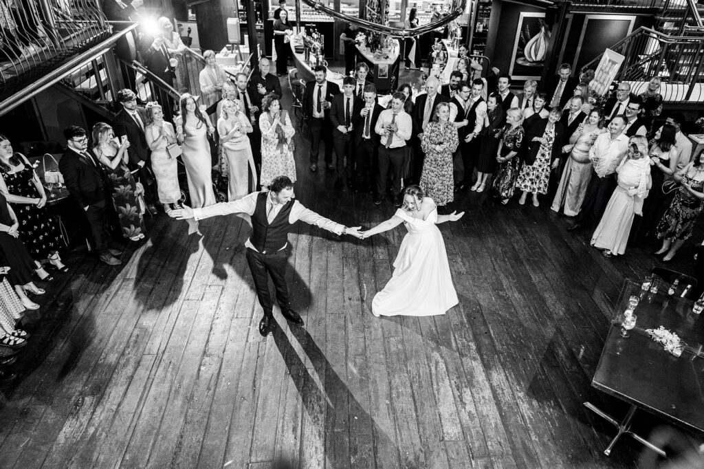A bride and groom dance in the center of a wooden floor, surrounded by a large group of guests watching and smiling in a warmly lit venue.