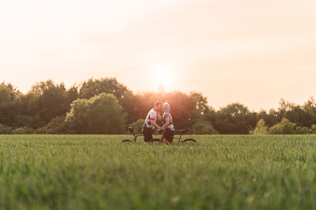Two people sit on a bicycle together in a green field at sunset, with trees in the background and warm sunlight creating a romantic atmosphere.