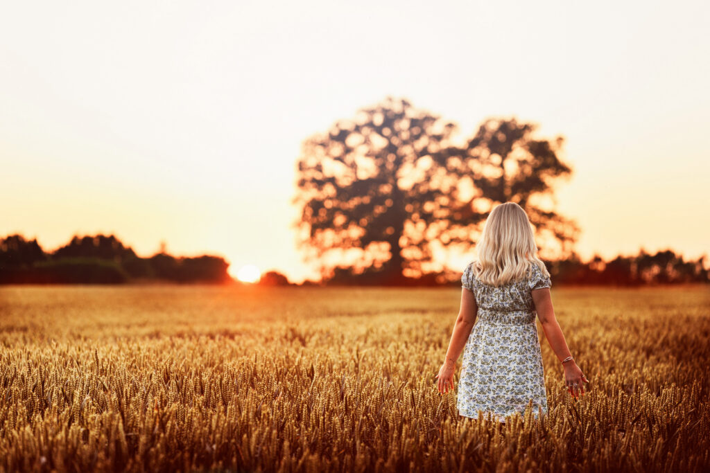 A woman with long blonde hair in a floral dress stands in a golden wheat field at sunset, facing away towards a large tree and the glowing sun on the horizon during her golden hour engagement shoot.