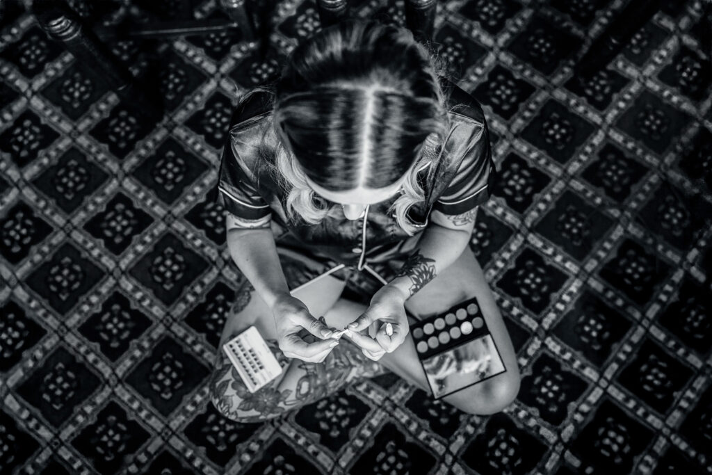 A woman with tattooed arms sits cross-legged on a patterned floor, seen from above, applying false eyelashes. Make-up palettes and packaging are scattered in front of her. The image is in black and white.