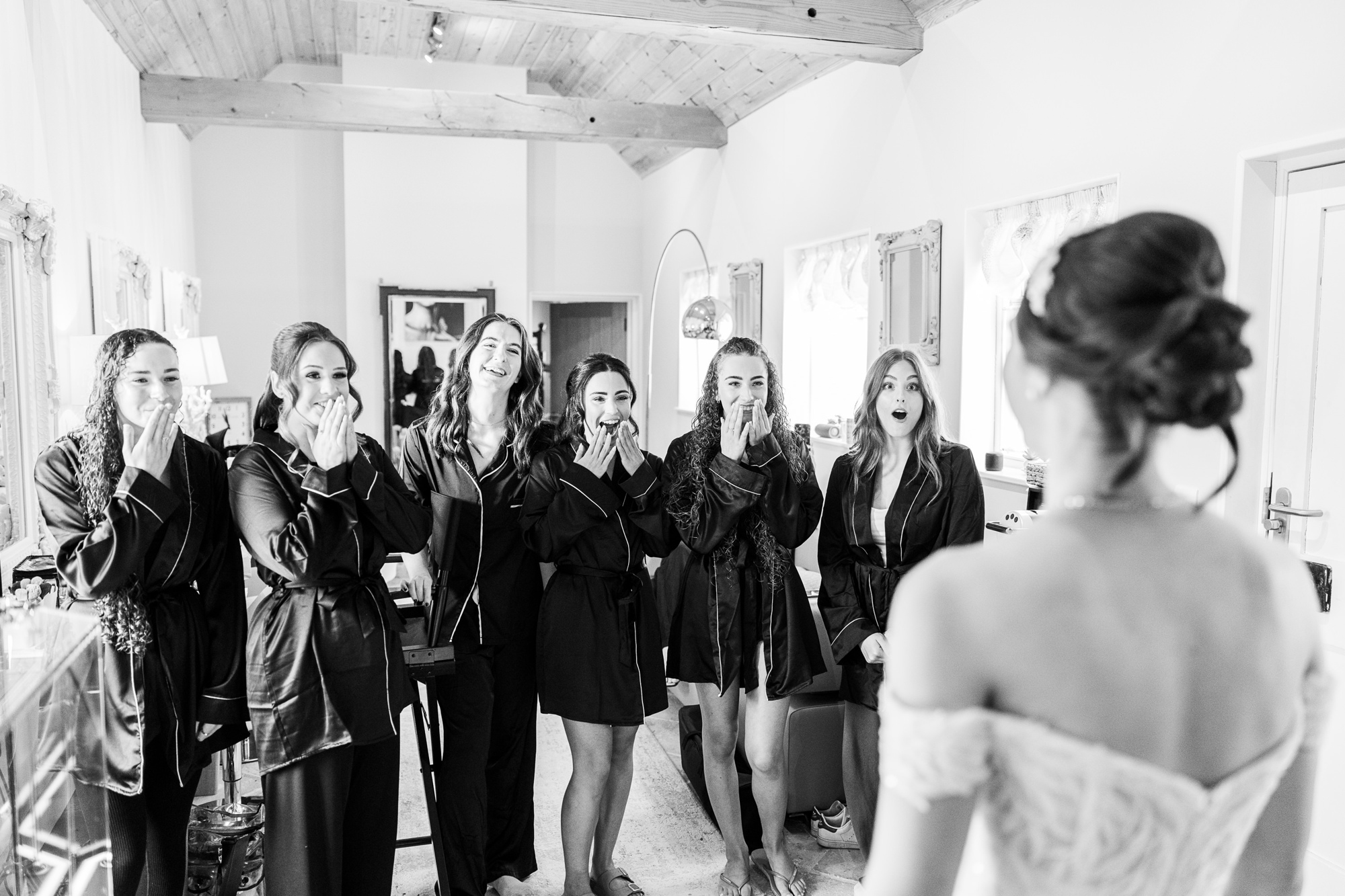3971624407 Six women in matching dressing gowns stand indoors, smiling and looking surprised with hands over their mouths, as a bride in a wedding dress faces them with her back to the camera. The scene is bright and joyful.