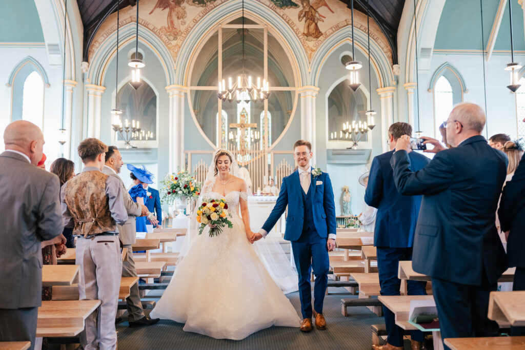 A bride and groom walk hand-in-hand down the aisle of a decorated church, smiling, as guests on both sides stand and watch, some taking photos. The church interior features arches, chandeliers, and stained glass.
