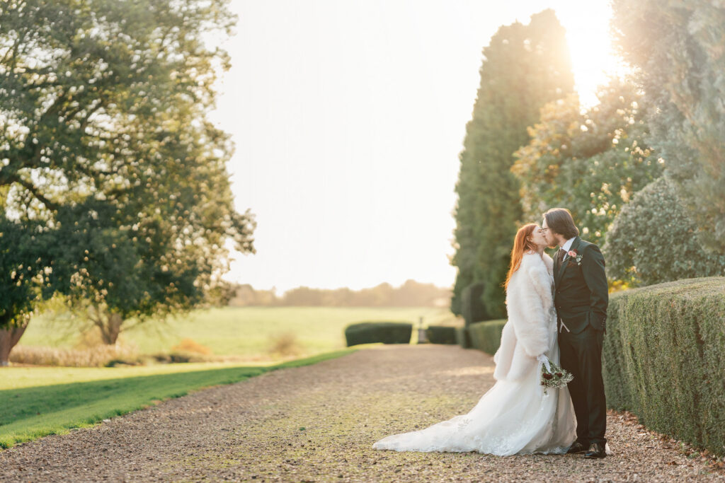 A bride and groom share a kiss on a sunlit garden path, surrounded by trees and greenery. The bride wears a white dress and fur shawl, while the groom is in a dark suit, holding her bouquet.