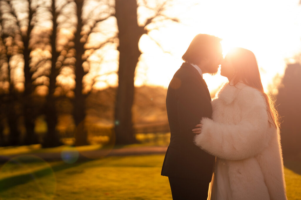 A couple stands close together at sunset in a park, with the woman wearing a white fur coat and the man in a dark suit. The sun creates a warm, glowing backlight, and trees are visible in the background.