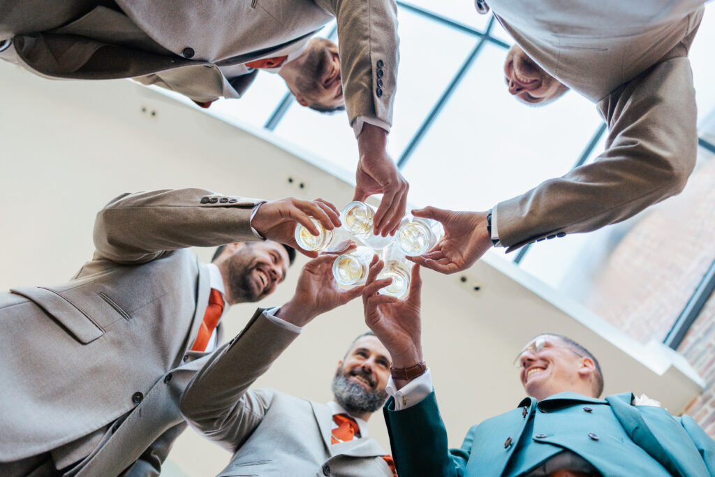 Five men in suits stand in a circle, raising glasses in a toast and smiling, viewed from below with a skylight above them.