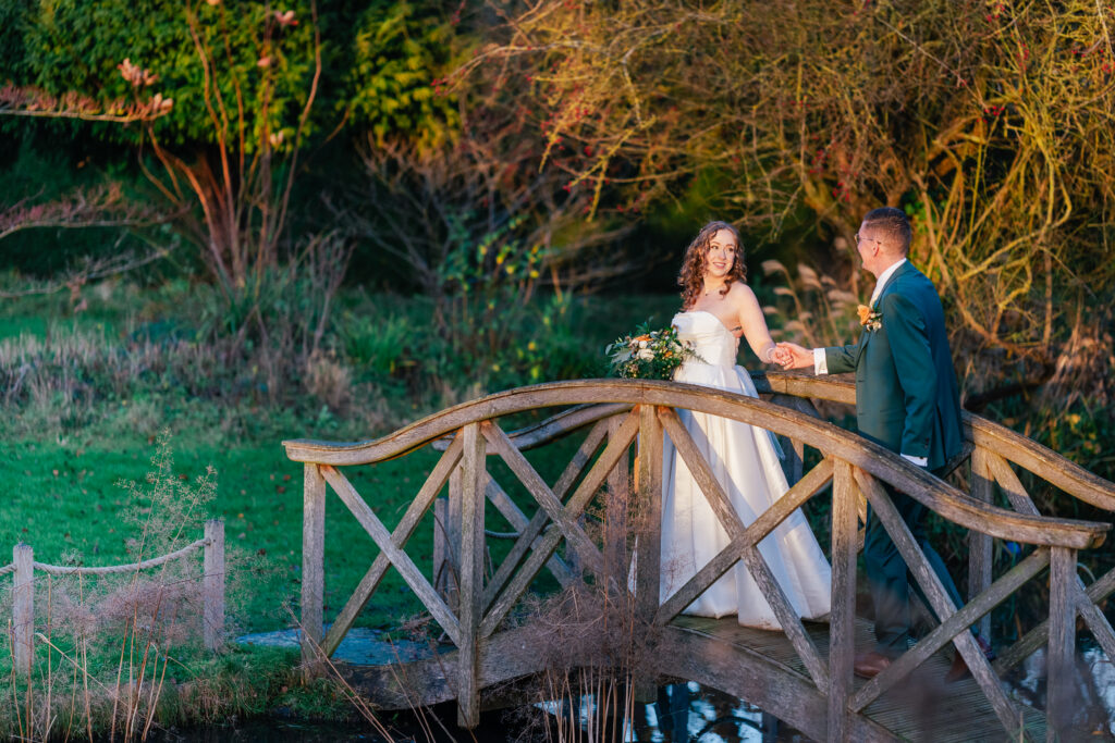 A bride in a white dress holding a bouquet and a groom in a green suit stand on a wooden bridge in a sunlit garden, holding hands and smiling at each other. Trees and greenery surround them.