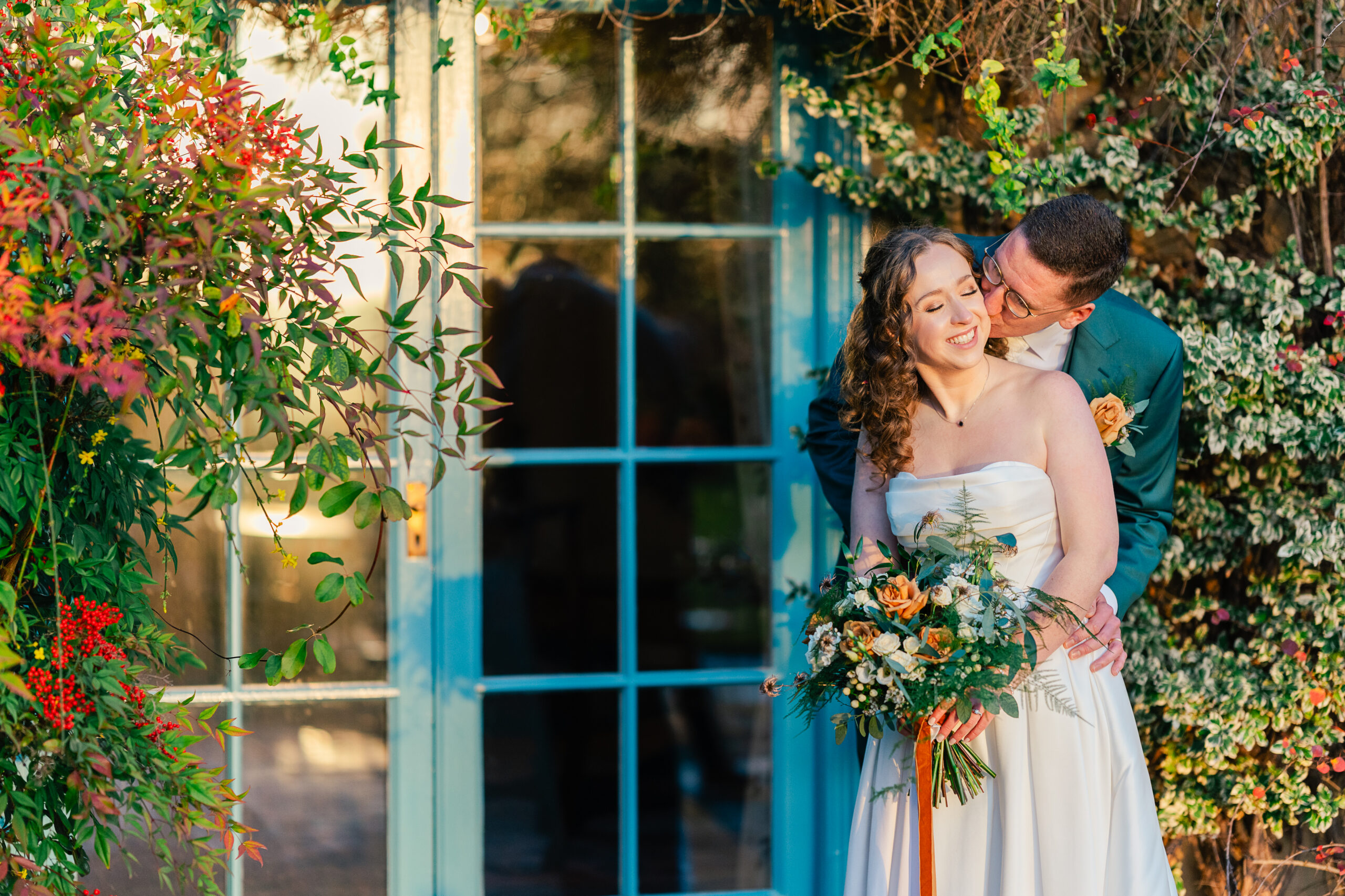 SouthFarm-5 A groom lovingly embraces and kisses his smiling bride, who holds a bunch of flowers, as they stand in front of a blue-framed glass door surrounded by lush greenery and colourful foliage.