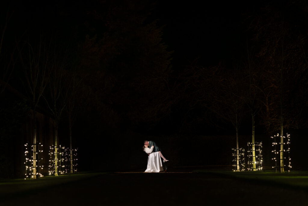 A couple dressed in wedding attire share a dramatic kiss at night, illuminated by soft white fairy lights wrapped around small trees lining a dark path. The background is mostly dark, focusing attention on the couple.